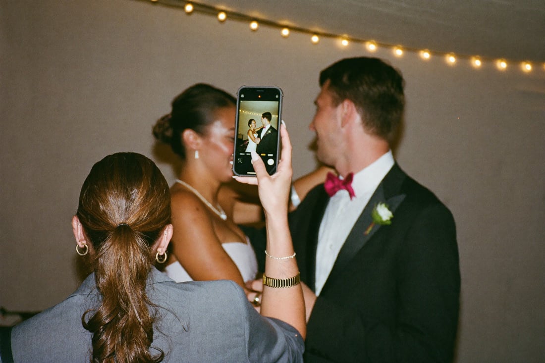 A guest holding up a phone to photograph the couple under string lights.